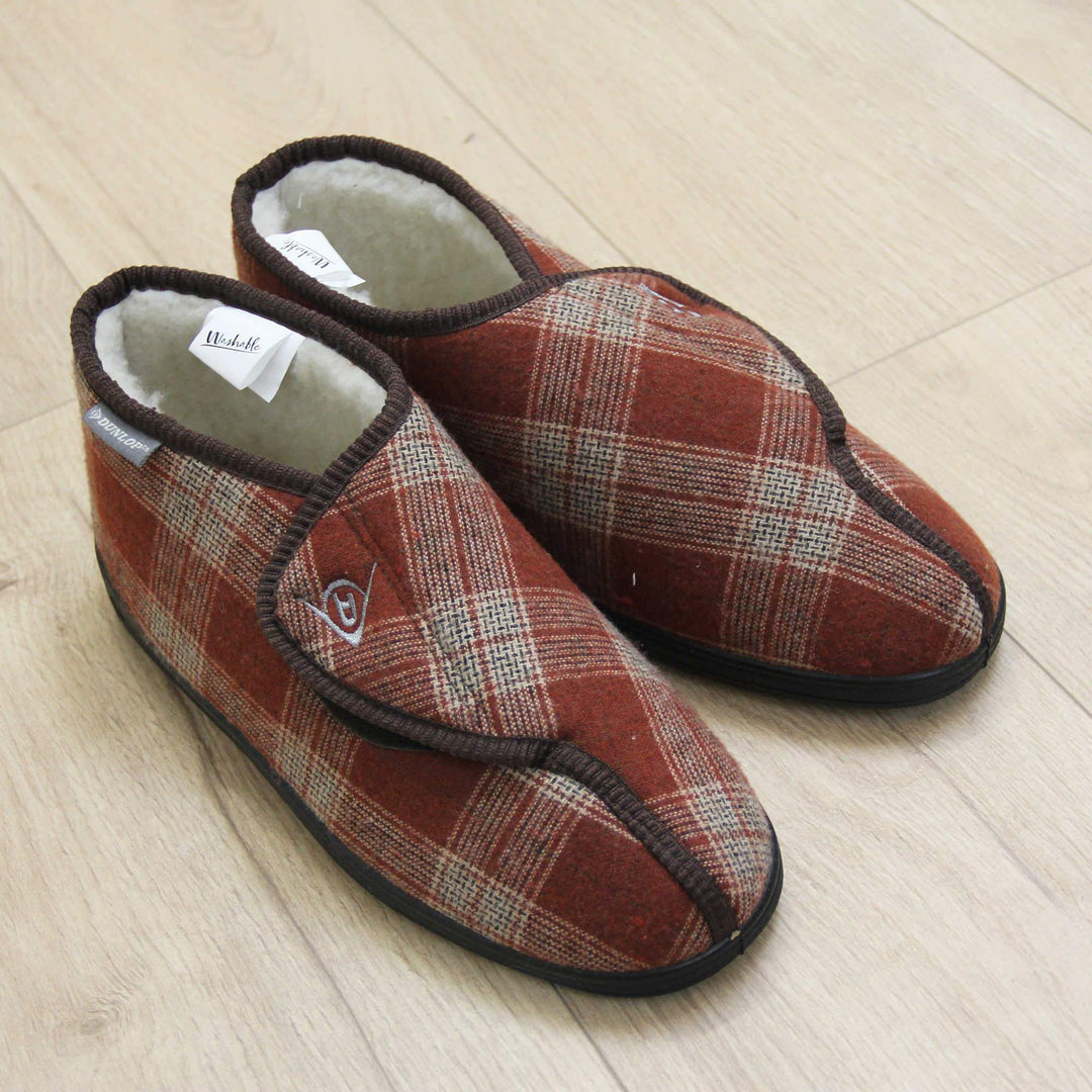 Orthopaedic boot slippers. Mens orthopaedic slippers in an ankle boot style. With a brown plaid upper and white fleece lining. With an adjustable touch close top with a grey Dunlop logo on. Small grey label to the outer side edge with Dunlop written on. Thick black outdoor sole. Lifestyle photo with both shoes together on a wood floor, taken from above