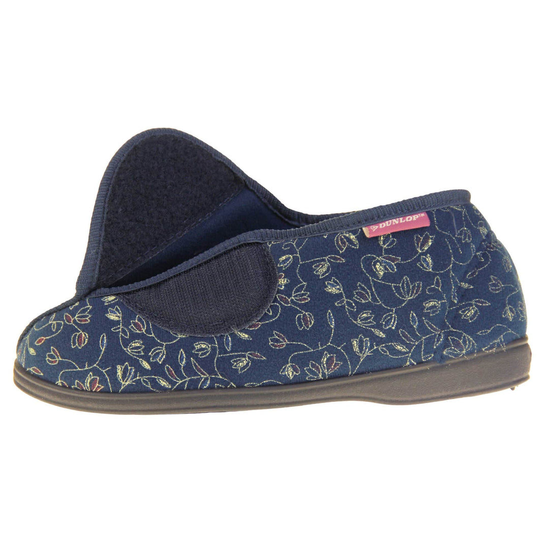 Orthopaedic slippers. Womens bootie style slipper with a navy blue textile upper with vine and flower embroidered design. Touch fasten tab to the top and blue textile lining. Firm black sole. Left foot from a side view with the touch fasten tab open.