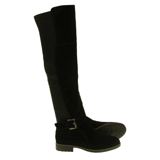 Over the knee flat suede boots. Tall boots that go above the knee in a black suede upper. Strap with buckle around the ankle. Black outsole with slight heel. Both feet from a side profile with the left foot on its side to show the sole.