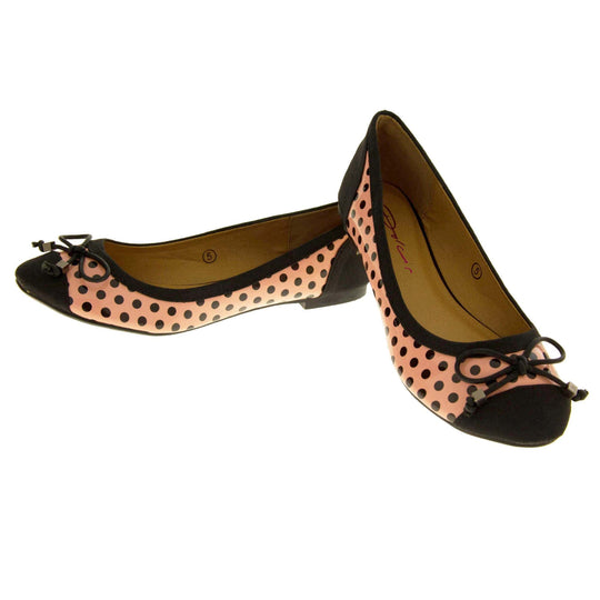 Polka dot flats. Womens ballerina style shoes with a glossy peach upper with black polka dots covering it. Black faux suede toes and heels and a black bow to the top. Nude faux-leather lining. Black sole with very slight heel. Both feet in a V shape with the back right foot resting on top of the back of the left foot.