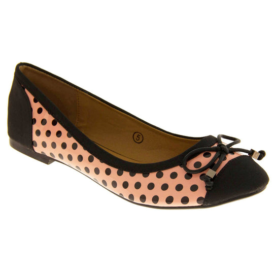 Polka dot flats. Womens ballerina style shoes with a glossy peach upper with black polka dots covering it. Black faux suede toes and heels and a black bow to the top. Nude faux-leather lining. Black sole with very slight heel. Right foot at an angle.