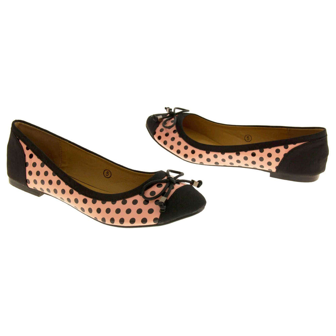 Polka dot flats. Womens ballerina style shoes with a glossy peach upper with black polka dots covering it. Black faux suede toes and heels and a black bow to the top. Nude faux-leather lining. Black sole with very slight heel. Both feet at an angle facing top to tail.