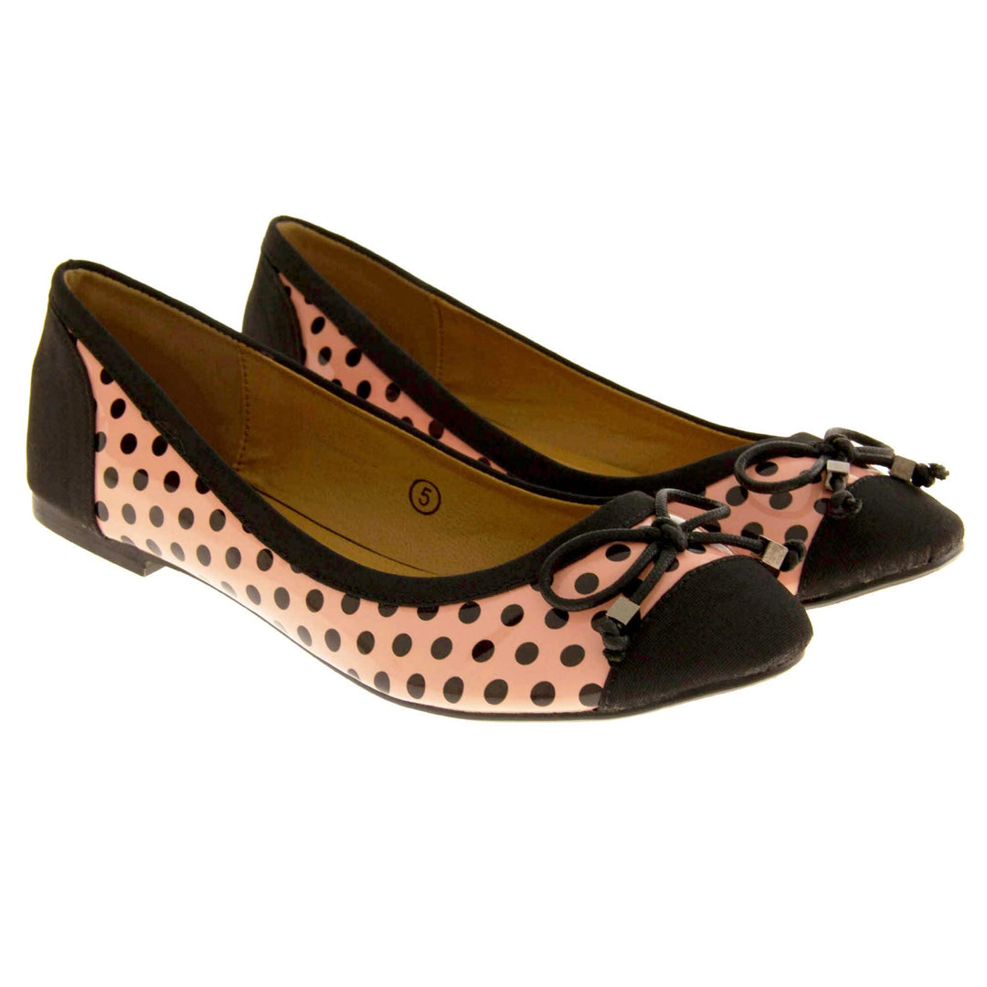 Polka dot flats. Womens ballerina style shoes with a glossy peach upper with black polka dots covering it. Black faux suede toes and heels and a black bow to the top. Nude faux-leather lining. Black sole with very slight heel. Both feet together at a slight angle.