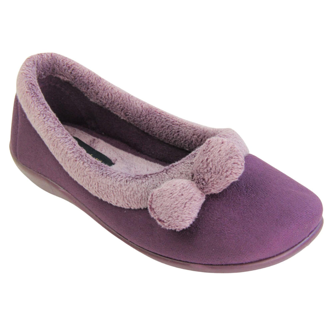 Pom pom slippers. Womens ballerina style slippers with purple velour uppers. Pale purple plush textile collar and two pom poms to the front of the shoe. Matching textile lining. Firm purple sole. Right foot at an angle.