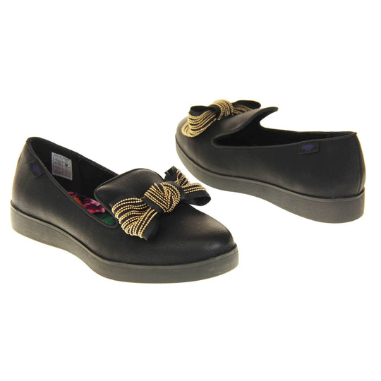 Rocket Dog slip on shoes. Loafer style shoes with a black faux leather upper. With a black and gold bow detail on the top of the foot. Bright floral patterned insole. Chunky black sole with slip resistant grip to the bottom.  Both feet at an angle facing top to tail.