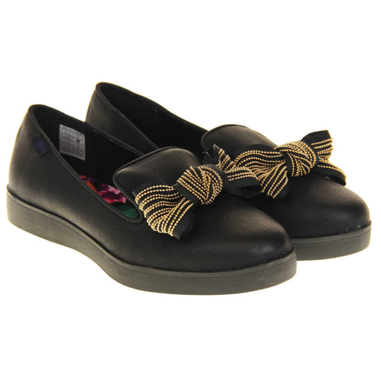 Rocket Dog slip on shoes. Loafer style shoes with a black faux leather upper. With a black and gold bow detail on the top of the foot. Bright floral patterned insole. Chunky black sole with slip resistant grip to the bottom. Both feet together at a slight angle.