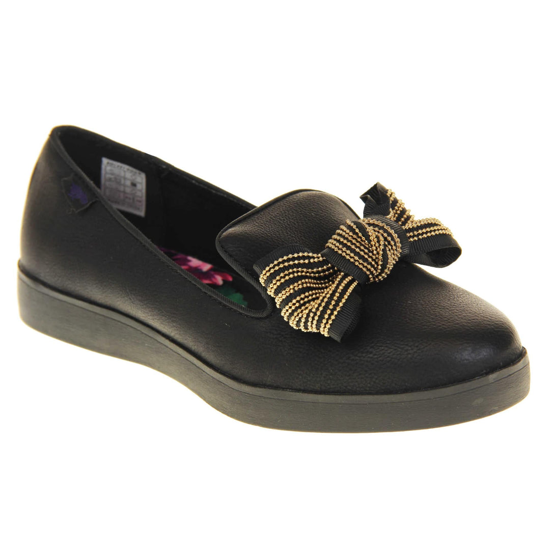 Rocket Dog slip on shoes. Loafer style shoes with a black faux leather upper. With a black and gold bow detail on the top of the foot. Bright floral patterned insole. Chunky black sole with slip resistant grip to the bottom. Right foot at an angle.