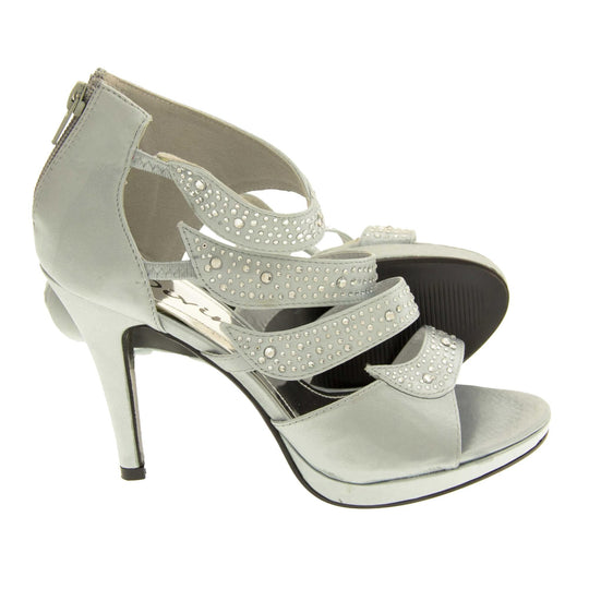Satin strappy heels. Womens high heels with open toes, 4 silver satin straps along the front covered in diamantes. Closed back in the same silver satin with zip up the back for putting them on. Metallic silver insoles. Silver satin stiletto heel and black sole.  Both feet from a side profile with the left foot on its side behind the the right foot to show the sole.