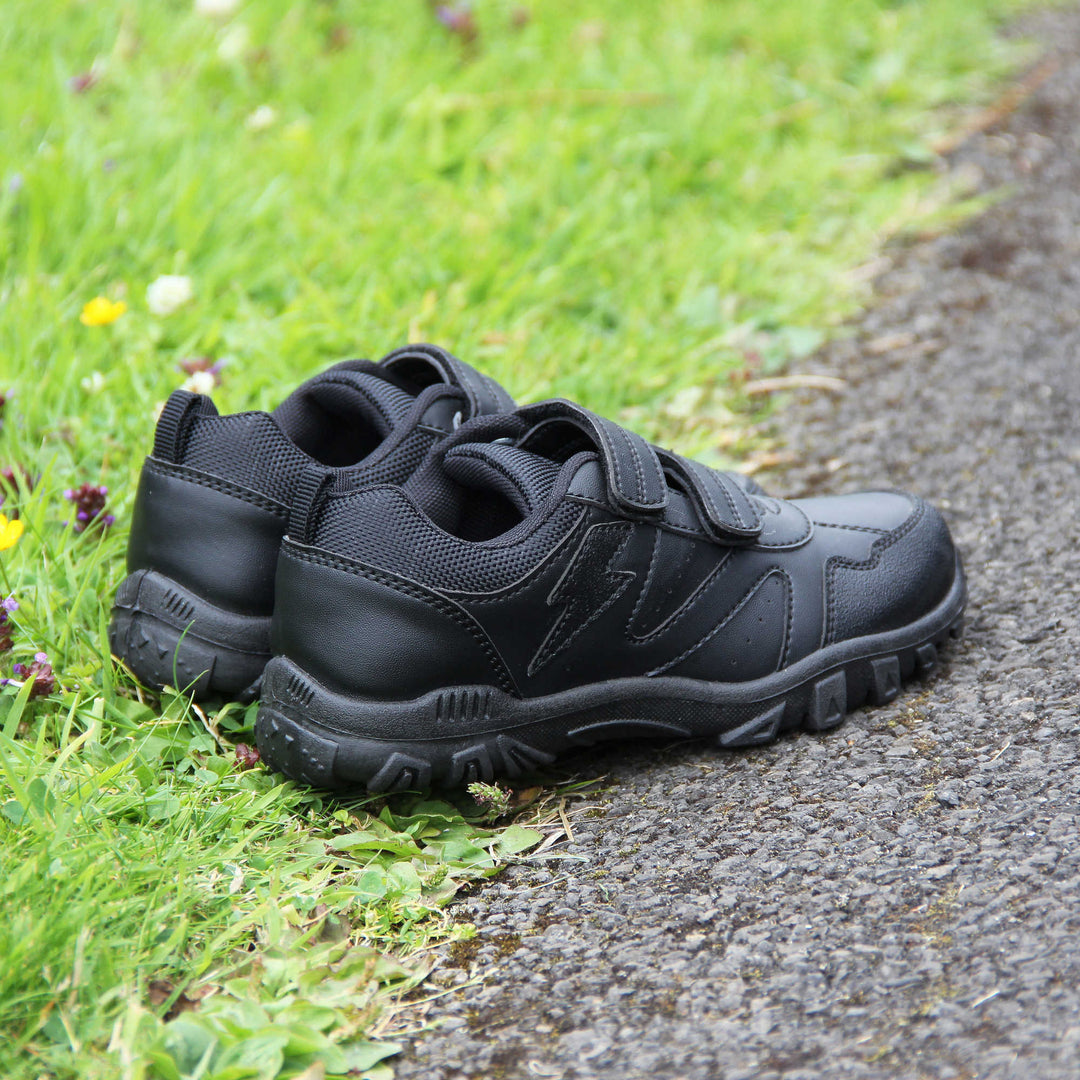 School trainers boy. Black faux leather trainers with zig zag detailing along the sides and a lightning bolt near the ankle. Two black touch fasten straps across the foot with a black textile lining. Chunky black soles.  Lifestyle photo with both shoes together at an angle half on some grass, half on a path.
