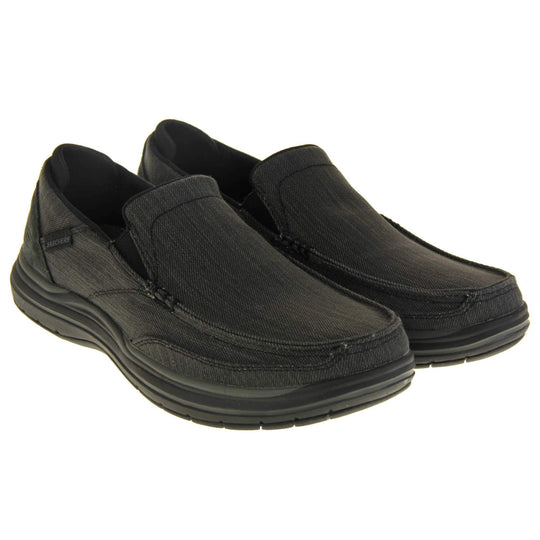 Skechers Slip On. Black loafer trainers with black denim upper with stitching detail. Black tag to the outside ankle with Skechers written on. The heel is a faux suede with S logo on it. Chunky black sole with grip to the bottom. Both feet at an angle next to each other.