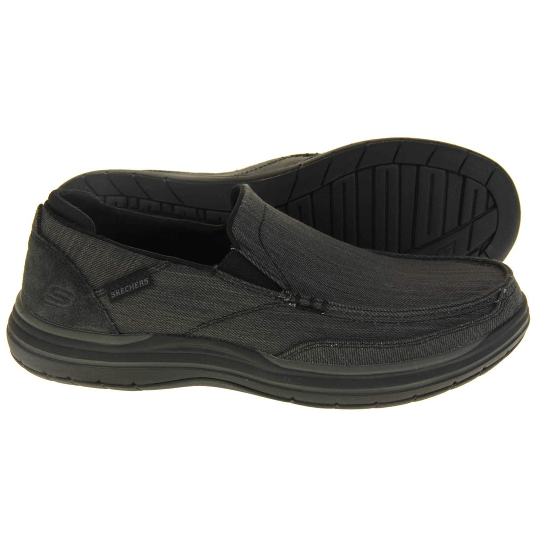 Skechers Slip On. Black loafer trainers with black denim upper with stitching detail. Black tag to the outside ankle with Skechers written on. The heel is a faux suede with S logo on it. Chunky black sole with grip to the bottom. Both feet from a side profile with the left foot on its side to show the sole.