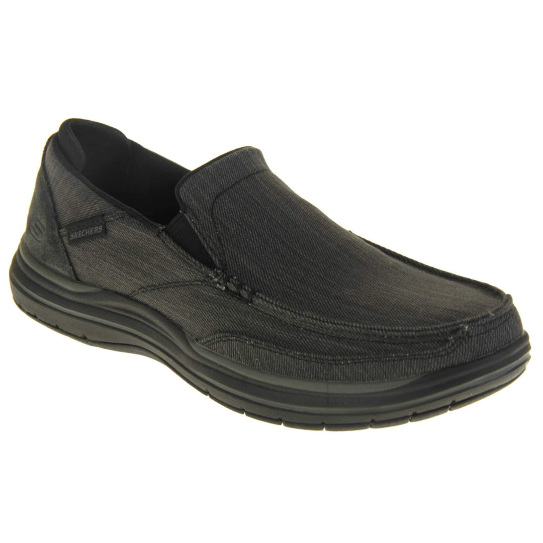 Skechers Slip On. Black loafer trainers with black denim upper with stitching detail. Black tag to the outside ankle with Skechers written on. The heel is a faux suede with S logo on it. Chunky black sole with grip to the bottom. Right foot at an angle.
