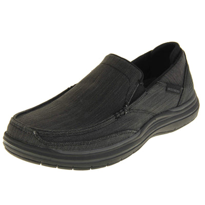 Skechers Slip On. Black loafer trainers with black denim upper with stitching detail. Black tag to the outside ankle with Skechers written on. The heel is a faux suede with S logo on it. Chunky black sole with grip to the bottom. Left foot at an angle.