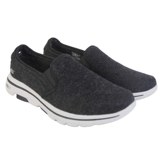 Skechers trainers. Loafer style shoes with a trainer sole. Black wool upper with elasticated panels by the tongue to give more flexibility and a better fit. Skechers S logo to the back. Chunky white sole with a black line running around it. Both feet together from an angle.