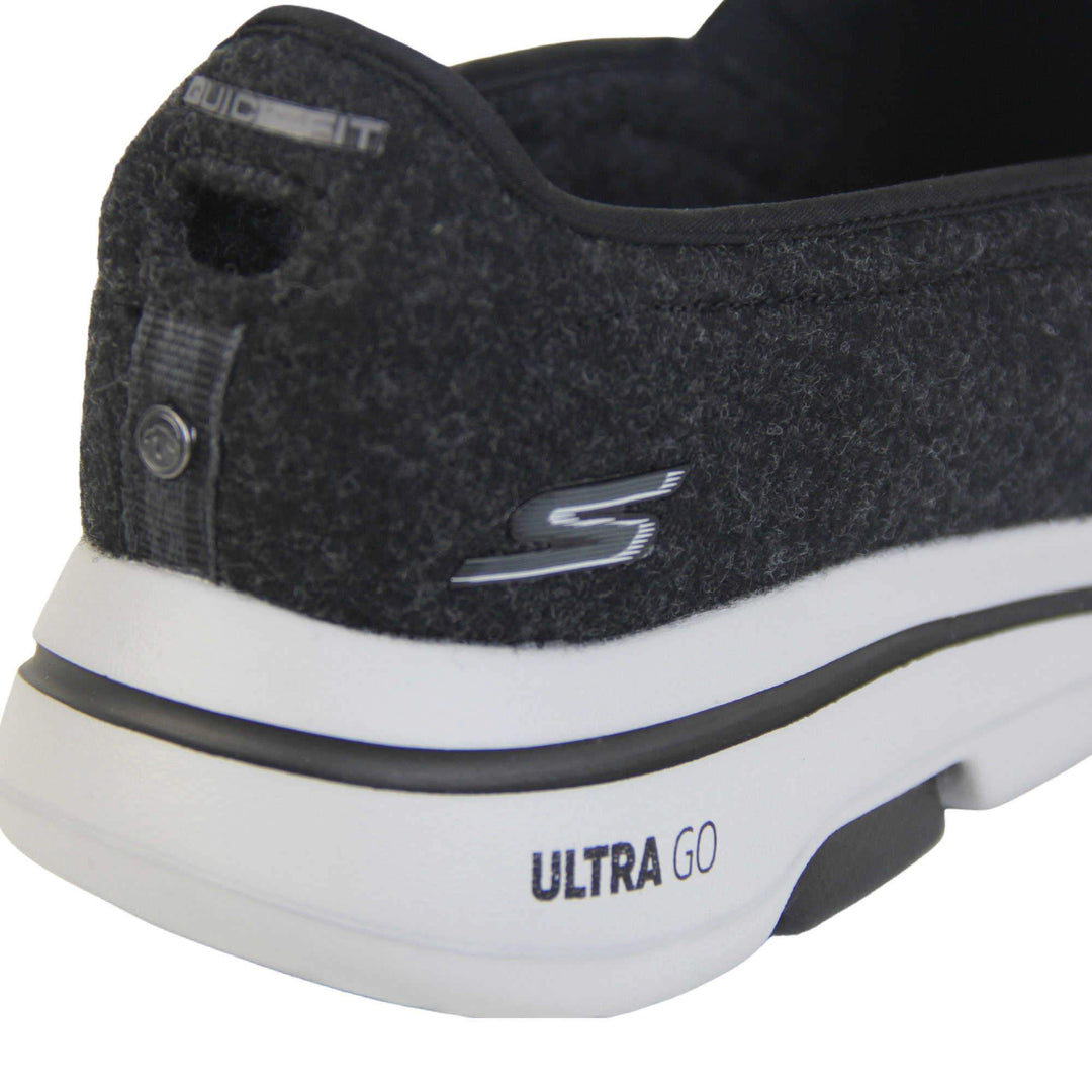 Skechers trainers. Loafer style shoes with a trainer sole. Black wool upper with elasticated panels by the tongue to give more flexibility and a better fit. Skechers S logo to the back. Chunky white sole with a black line running around it. Close up of the shoe to show the back of it and Skechers detail to the back.