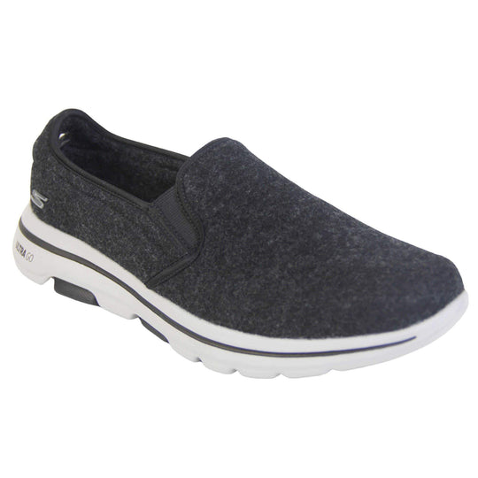 Skechers trainers. Loafer style shoes with a trainer sole. Black wool upper with elasticated panels by the tongue to give more flexibility and a better fit. Skechers S logo to the back. Chunky white sole with a black line running around it. Right foot at an angle.