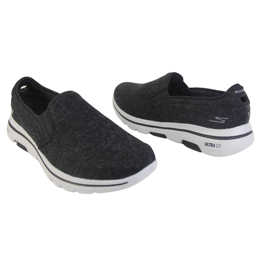 Skechers trainers. Loafer style shoes with a trainer sole. Black wool upper with elasticated panels by the tongue to give more flexibility and a better fit. Skechers S logo to the back. Chunky white sole with a black line running around it. Both feet from a slight angle facing top to tail.