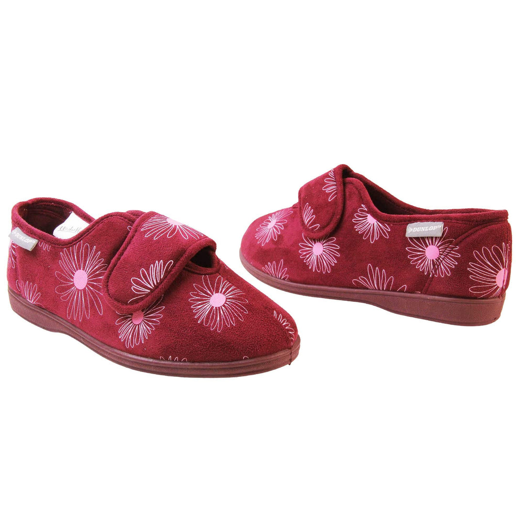 Slippers elderly women. Womens full back slipper. With a plum coloured upper with white daisy design and pink for the middle of the flowers. Touch fasten strap over the top of the foot to adjust the fit. Plum textile lining and firm plum sole. Both feet at an angle, facing top to tail.