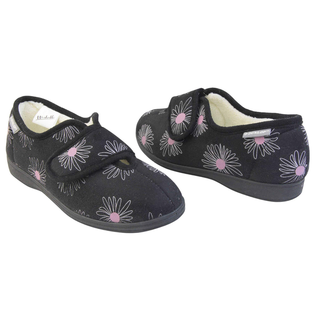 Slippers for elderly with swollen feet. Womens full-back slipper. Black upper with a white daisy design and pink for the middle of the flowers. Touch fasten strap over the top of the foot to adjust the fit. Cream fleece lining and firm black sole. Both feet at an angle, facing top to tail.