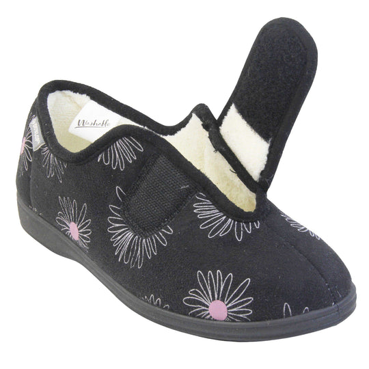 Slippers for elderly with swollen feet. Womens full-back slipper. Black upper with a white daisy design and pink for the middle of the flowers. Touch fasten strap over the top of the foot to adjust the fit. Cream fleece lining and firm black sole. Left foot at an angle with the touch fasten strap open to show the adjustability.
