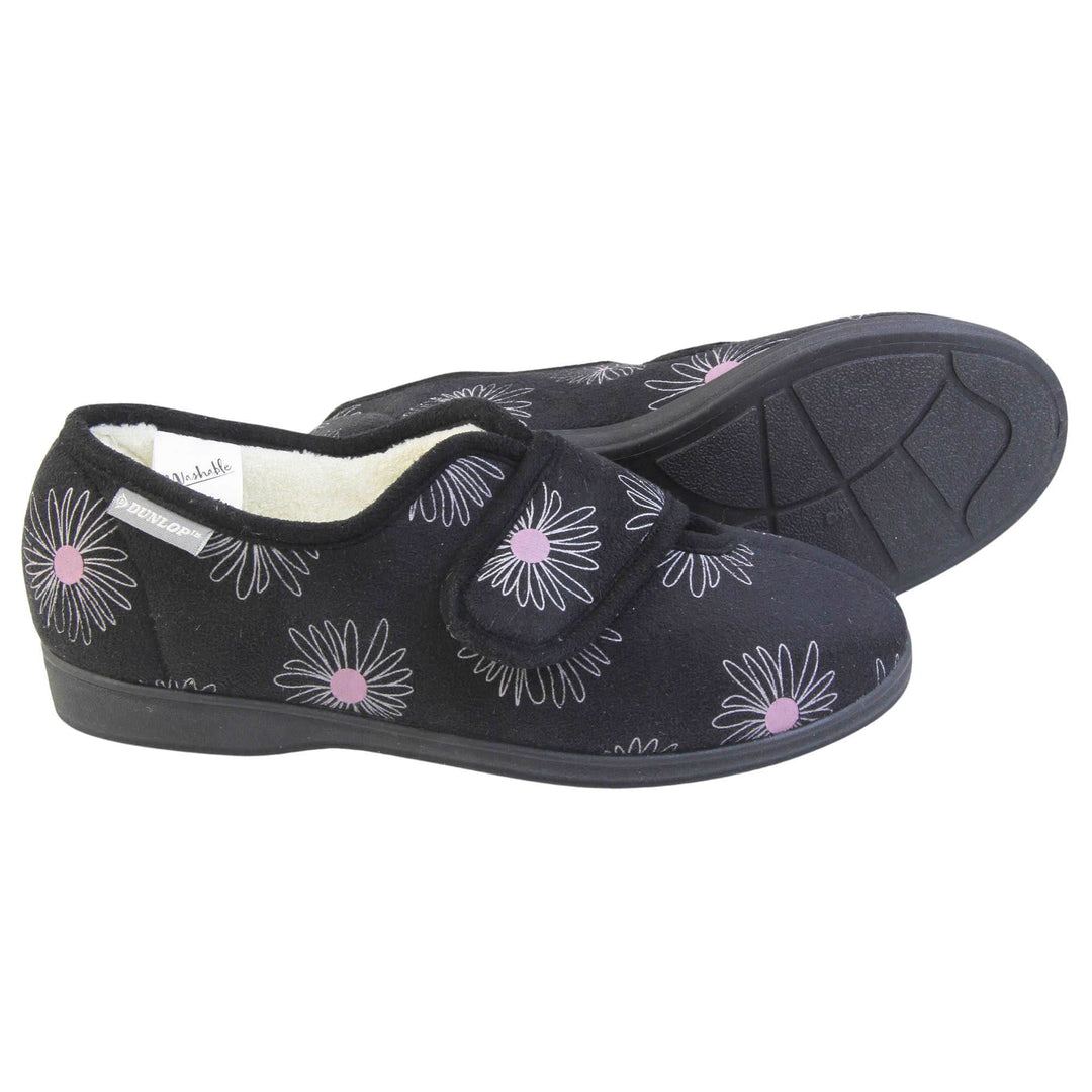 Slippers for elderly with swollen feet. Womens full-back slipper. Black upper with a white daisy design and pink for the middle of the flowers. Touch fasten strap over the top of the foot to adjust the fit. Cream fleece lining and firm black sole. Both feet from a side profile with the left foot on its side behind the right foot to show the sole.