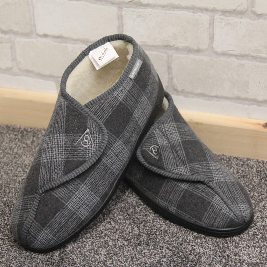 Orthopaedic slippers. Mens orthopaedic slippers in an ankle boot style. With a grey plaid upper and white fleece lining. With an adjustable touch close top with a grey Dunlop logo on. Small grey label to the outer side edge with Dunlop written on. Thick black outdoor sole. Lifestyle photo with both feet in a V shape with the left foot heel stacked on top of the right. On a grey carpet with grey brick wall behind a pale wood skirting board.