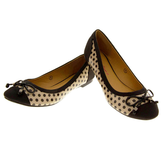 Spotty Shoes. Womens ballerina style shoes with a glossy cream upper with black polka dots covering it. Black faux suede toes and heels and a black bow to the top. Nude faux-leather  lining. Black sole with very slight heel. Both feet in a V shape with the back right foot resting on top of the back of the left foot.