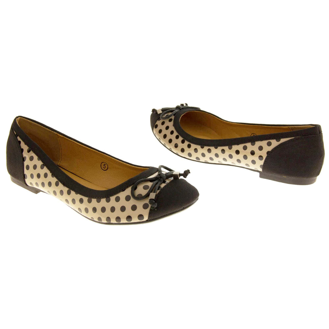 Spotty Shoes. Womens ballerina style shoes with a glossy cream upper with black polka dots covering it. Black faux suede toes and heels and a black bow to the top. Nude faux-leather  lining. Black sole with very slight heel. Both feet at an angle facing top to tail.