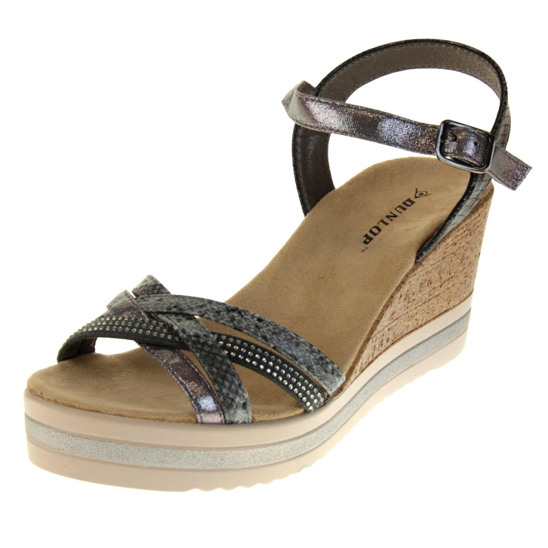 Strappy wedge sandals. Pewter coloured, faux leather strappy upper. The toe straps. are a mix between plain, faux snake skin and studded. Ankle strap is faux snakeskin. Nude faux suede cushioned insoles with black Dunlop branding. Cork wedge heel and platform sole made up of nude and white contrasting strips. Left foot at an angle.