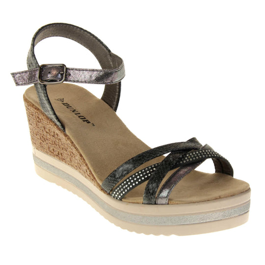 Strappy wedge sandals. Pewter coloured, faux leather strappy upper. The toe straps. are a mix between plain, faux snake skin and studded. Ankle strap is faux snakeskin. Nude faux suede cushioned insoles with black Dunlop branding. Cork wedge heel and platform sole made up of nude and white contrasting strips. Right foot at an angle.