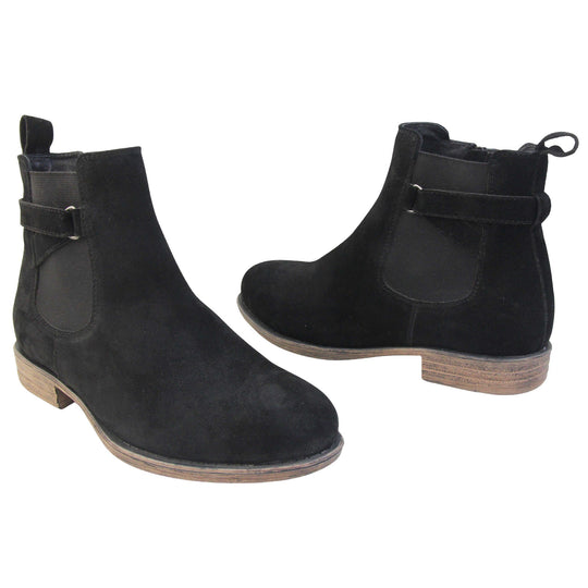 Suede leather ankle boots. Chelsea boot style with a black suede upper. Black elasticated panels at the ankles with straps in a zigzag across the panel. Black loop at the back to help pull them on. Brown sole with a slight heel. Both feet from a slight angle facing top to tail.
