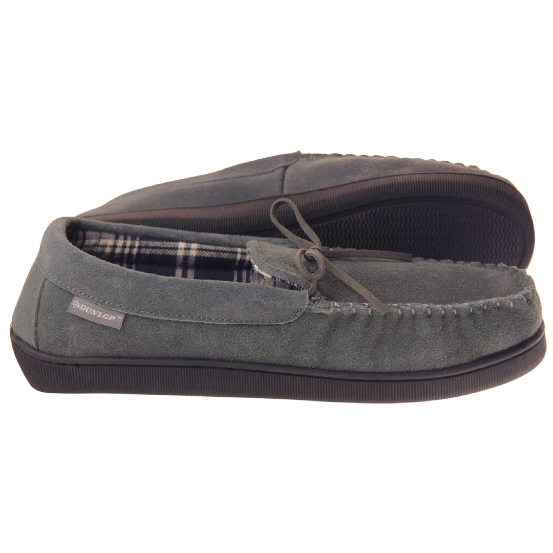 Suede moccasin slippers. Moccasin style slipper with grey suede upper and leather bow to the top. Grey Dunlop label to the outside. Grey and white plaid textile lining. Black rubber sole. Both feet from a side profile with the left foot on its side to show the sole.