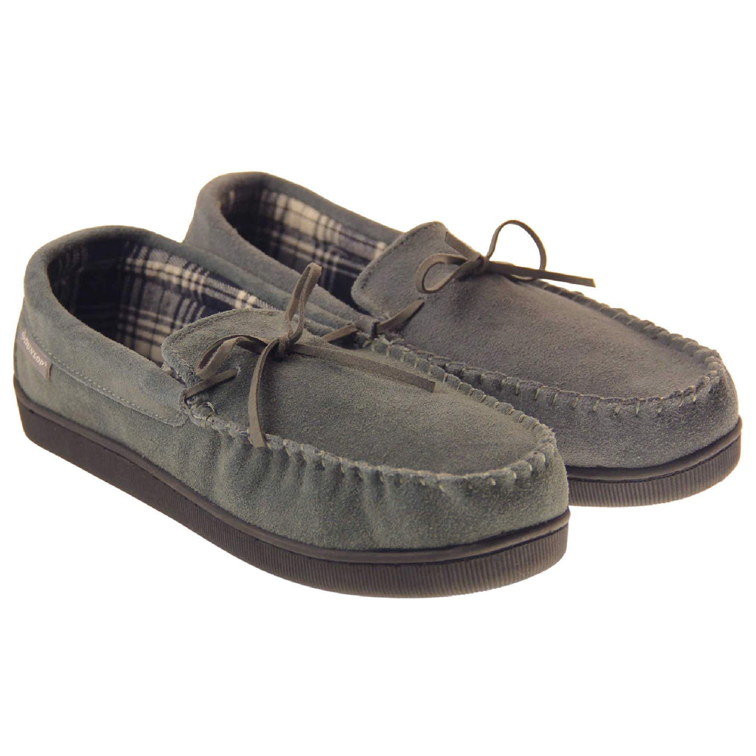 Suede moccasin slippers. Moccasin style slipper with grey suede upper and leather bow to the top. Grey Dunlop label to the outside. Grey and white plaid textile lining. Black rubber sole. Both feet together at an angle.