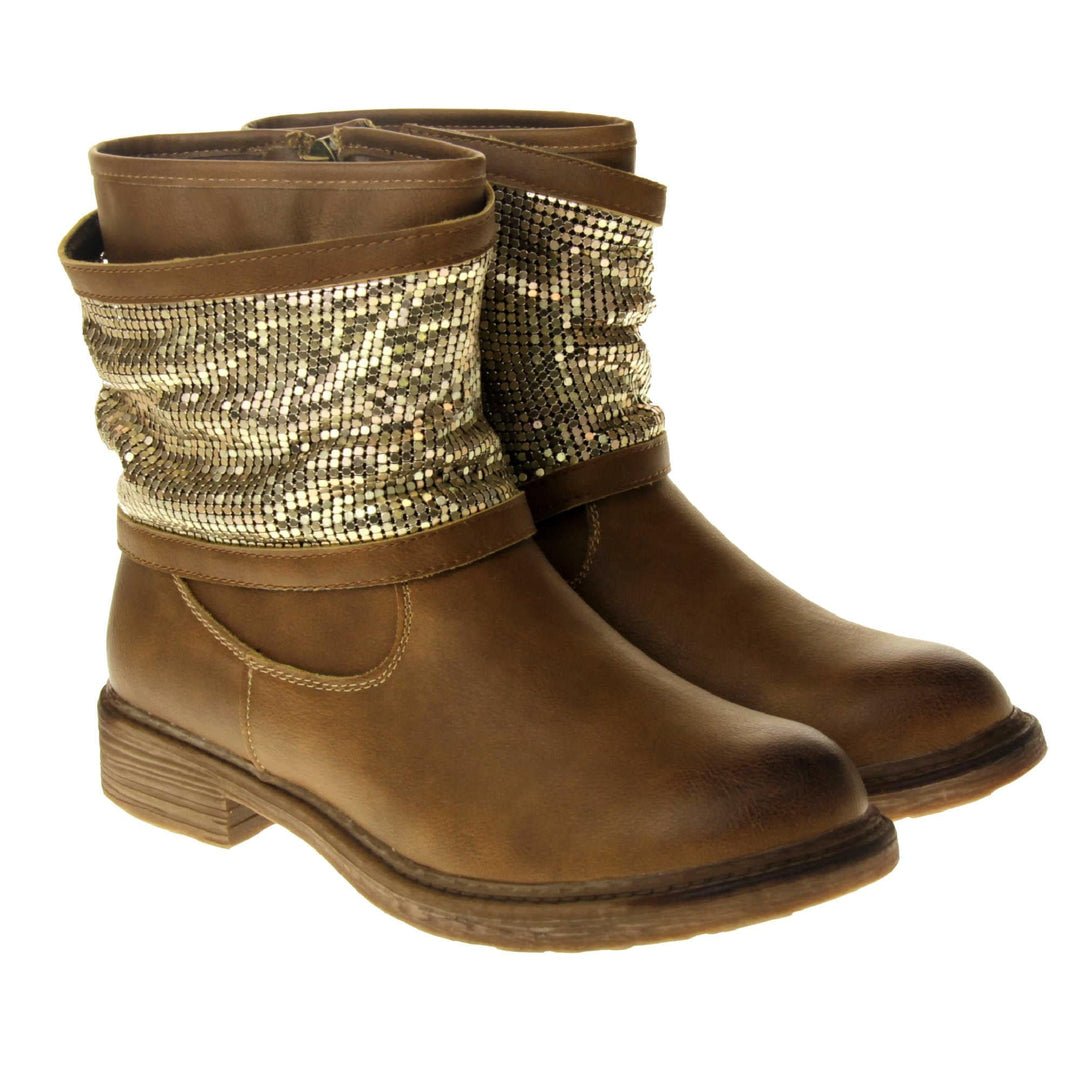 Tan sparkle ankle boots women. Biker style ankle boot with a tan faux leather upper. A thick band of shiny gold chainmail runs around the ankle. Zip fastening down the inside of the boot. Brown sole with a slight heel. Both feet together from an angle.