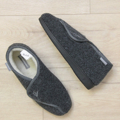 Orthopaedic slippers. Mens orthopaedic slippers in an ankle boot style. With a grey felt upper and white fleece lining. With an adjustable touch close top with a grey Dunlop logo on. Small grey label to the outer side edge with Dunlop written on. Thick black outdoor sole. Lifestyle photo from above of both slippers top to tale on a wood floor with the left foot on its side.