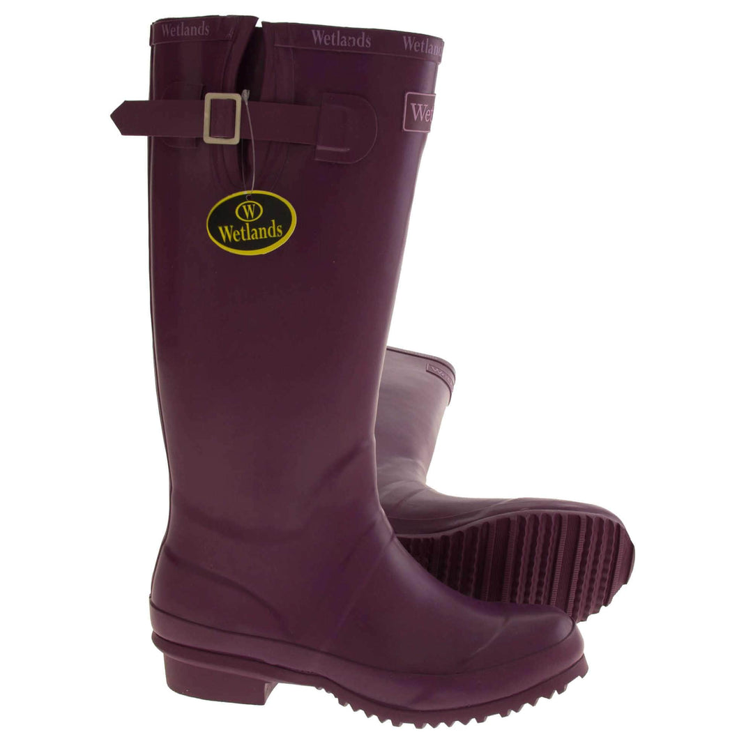 Womens Wellies. Purple rubber waterproof wellington boots. Just below knee height. With a small heel and deep tread to the sole. Pink Wetlands Brand to the front of the boot near the top. Wetlands written in Lilac numerous times around the rim of the boot. Side strap with buckle to adjust the calf width. Textile lining to the inside of the boot. Both feet from a side profile but with the left foot on it side to show you the sole.