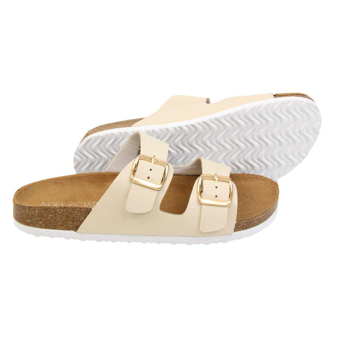 White double strap sandals. Womens dual strap slip on sandals. With a white synthetic leather upper with a gold buckle on each strap. Brown faux suede insole with a moulded footbed. Cork effect outsole with white base with grip to the bottom. Both feet from a side profile with the left foot on its side behind the the right foot to show the sole.