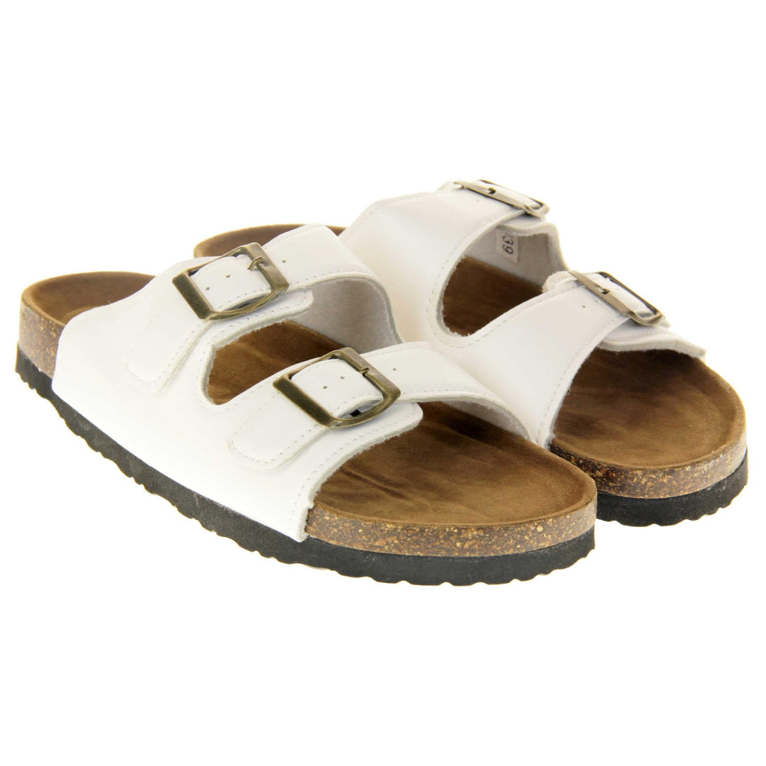White flat sandals womens. Ladies dual strap slip on sandals. With a white synthetic leather upper with a gold buckle on each strap. Brown faux suede insole with a moulded footbed. Cork effect outsole with black base with grip to the bottom. Both feet together at a slight angle.