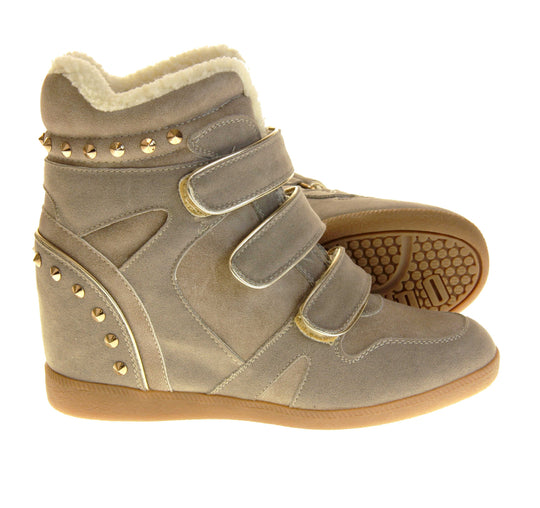 Women's wedge ankle boot. Ankle boot in a sneaker style with a grey faux suede upper. Wedge heel is hidden so the boots look flat. Three touch fasten straps across the front for fastening. Cream faux fur lining and trim. Gold studs around the collar and heel. Beige sole with good grip to the bottom. Both feet from a side profile with the left foot on its side to show the sole.