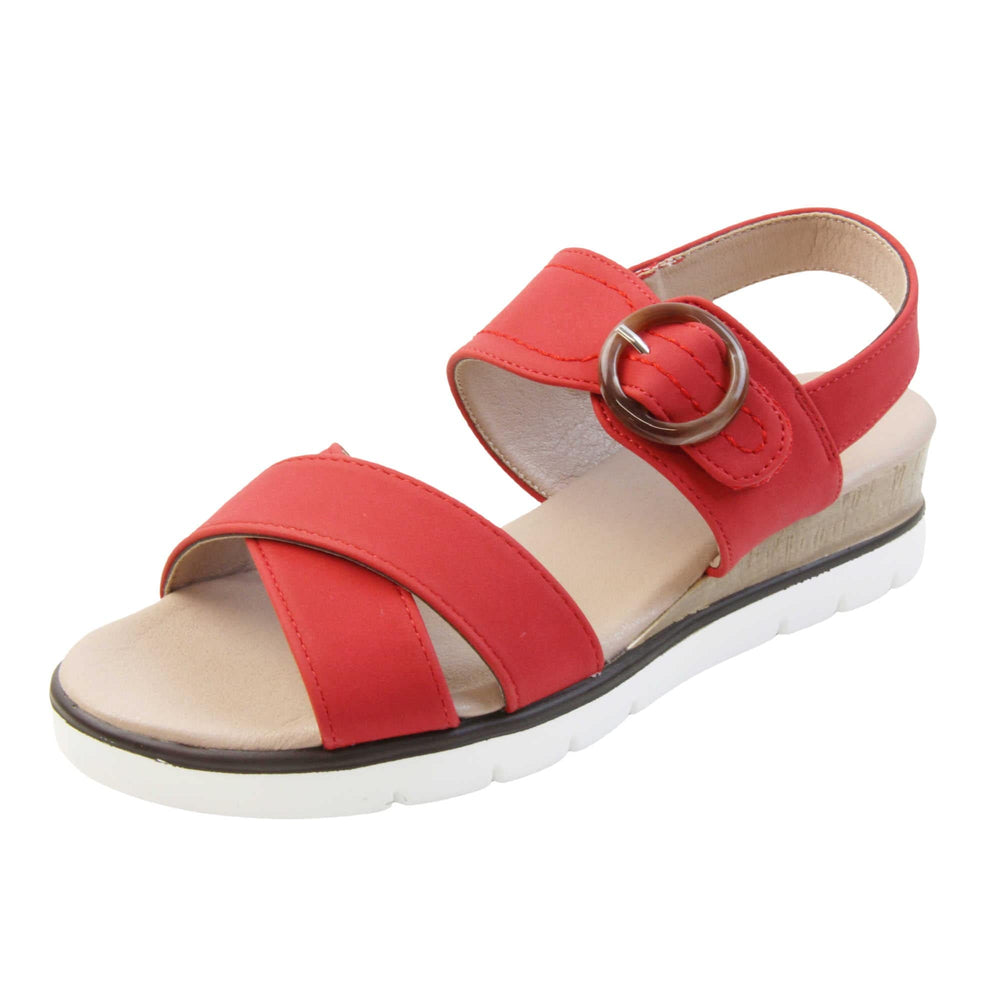 Womens wedge sandals. Classic womens strappy sandals with red textile straps. Dual toe straps that cross over each other. The ankle strap is touch fasten but has a brown buckle detail to look like a buckle fastening. Beige faux leather memory foam insoles. Small wedge heel in cork effect. White outsole with a black rim around the top. Left foot at an angle.