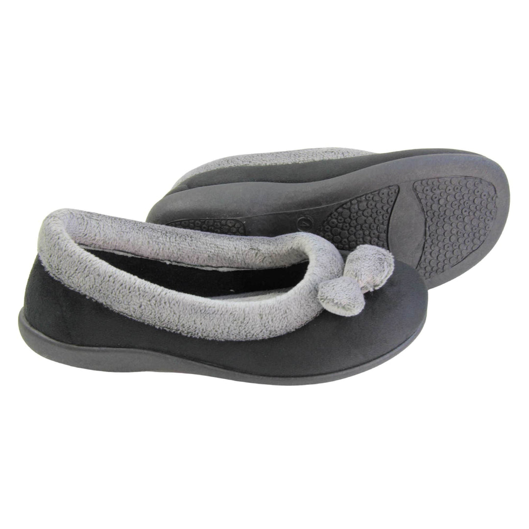 Womens ballerina slippers. Womens ballerina style slippers with black velour uppers. Grey, plush textile collar and two pom poms to the front of the shoe. Matching textile lining. Firm black sole. Both feet from a side profile with the left foot on its side behind the the right foot to show the sole.
