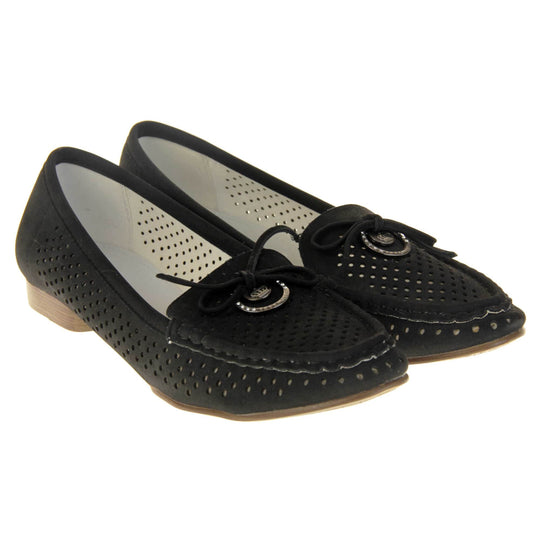 Womens black boat shoes. loafer style shoes with a black faux suede upper. Cut out dots along the sides and top of the shoe. Bow detail with metal hoop to the tongue of the shoe. Cream lining. Brown sole with slight heel. Both feet together at a slight angle.