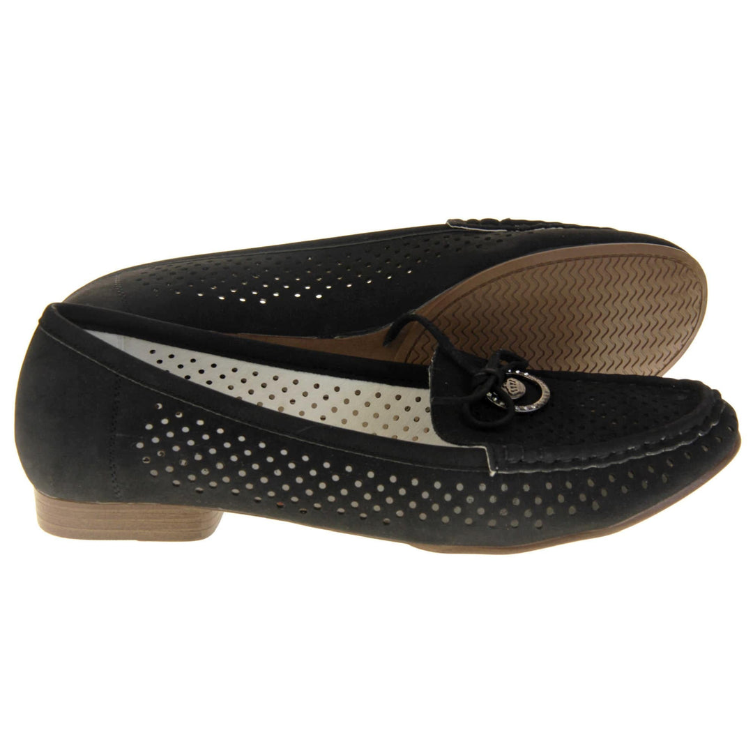 Womens black boat shoes. loafer style shoes with a black faux suede upper. Cut out dots along the sides and top of the shoe. Bow detail with metal hoop to the tongue of the shoe. Cream lining. Brown sole with slight heel. Both feet from a side profile with the left foot on its side behind the the right foot to show the sole.