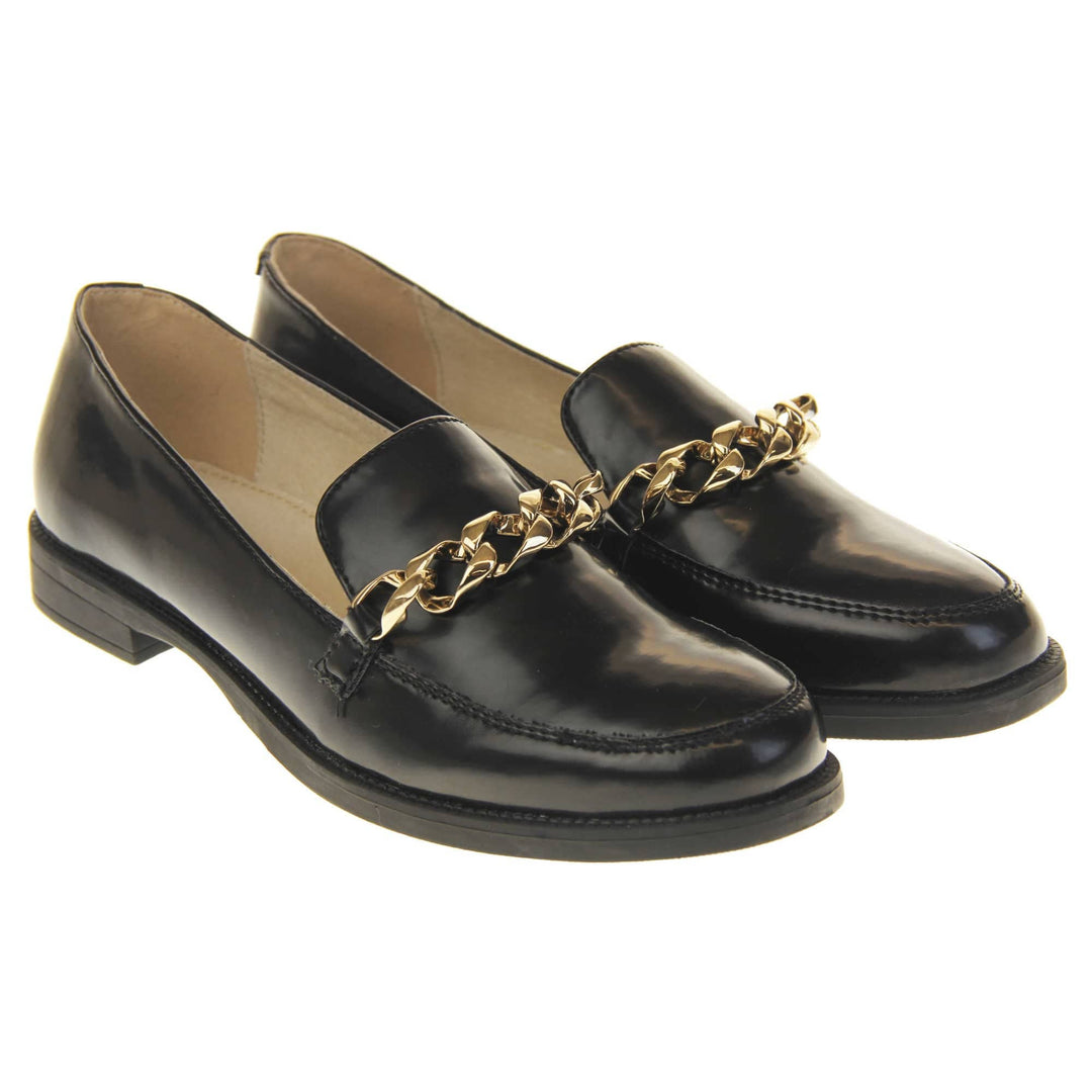Womens black brogues. Loafer shoes in a Brogue style with a black faux leather upper. Gold chain detail over the tongue. Real leather lining. Black sole with a slight heel. Both feet together at a slight angle.