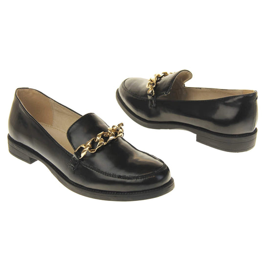 Womens black brogues. Loafer shoes in a Brogue style with a black faux leather upper. Gold chain detail over the tongue. Real leather lining. Black sole with a slight heel. Both feet at an angle facing top to tail.