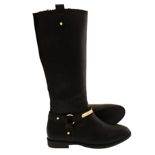 Womens black leather knee high boots. Knee high style boots with a black faux leather upper. A Tri strap going around the ankle and down to the bottom of the boot joined by the ankle with a good loop. Gold plate to the front of the ankle strap. Zip up the inside of the boot. Faux leather loop on the top of the outside of the boot to help with pulling it on. Both feet from a side profile with the left foot on its side to show the sole.