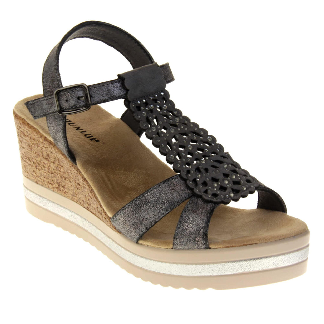 Womens black wedges. Strappy sandals with black faux leather upper. The central t bar has cut out detailing with diamantes. Nude faux suede cushioned insoles with black Dunlop branding. Cork wedge heel and platform sole made up of nude and white contrasting strips. Right foot at an angle.