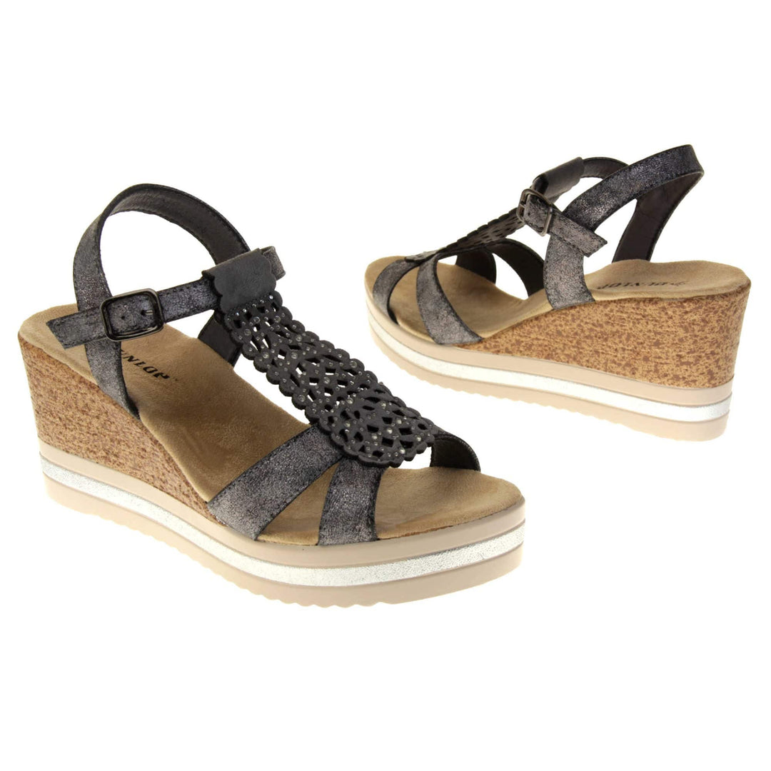 Womens black wedges. Strappy sandals with black faux leather upper. The central t bar has cut out detailing with diamantes. Nude faux suede cushioned insoles with black Dunlop branding. Cork wedge heel and platform sole made up of nude and white contrasting strips. Both feet at an angle facing top to tail.