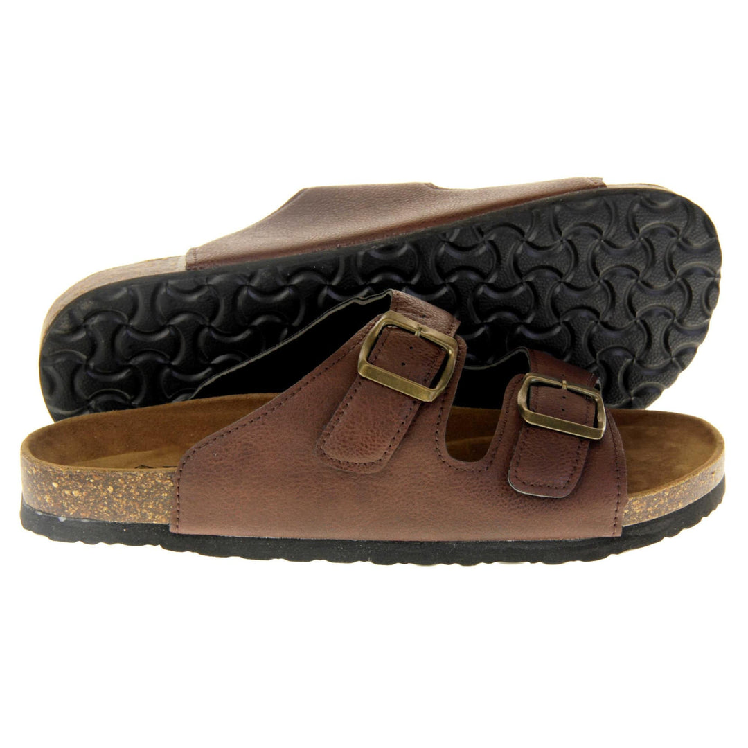 Womens brown slide sandals. Ladies dual strap slip on sandals. With a brown synthetic leather upper with a gold buckle on each strap. Brown faux suede insole with a moulded footbed. Cork effect outsole with black base with grip to the bottom. Both feet from a side profile with the left foot on its side behind the the right foot to show the sole.