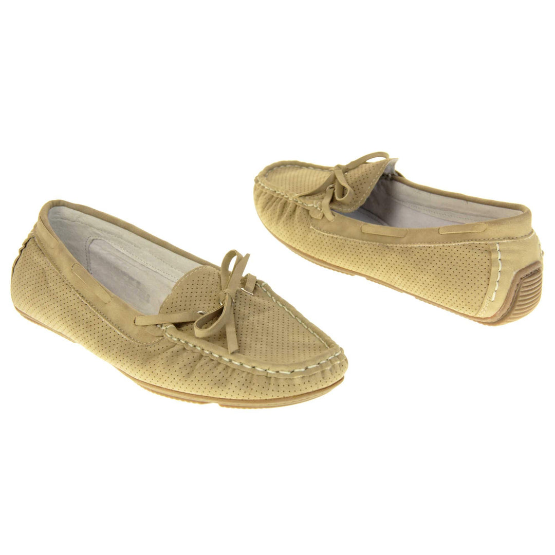 Women's deck shoes. Loafer style boat shoes with a beige faux leather upper and tiny dot cut-outs. Beige lace detail running around the outside of the collar. White stitching detailing around the top of the shoe. Cream leather lining and beige sole. Both feet at an angle facing top to tail.